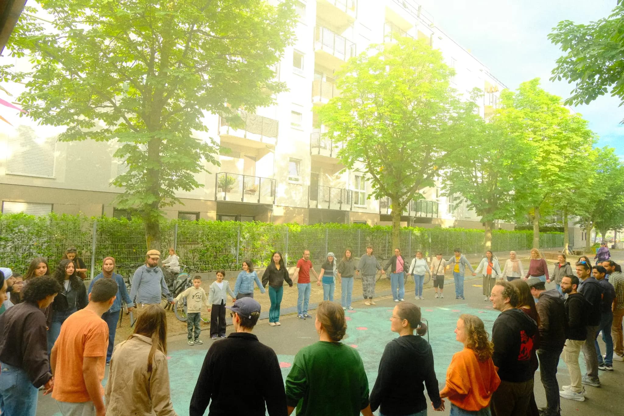 A community group holding hands in a large Dabke circle outdoors beneath trees.